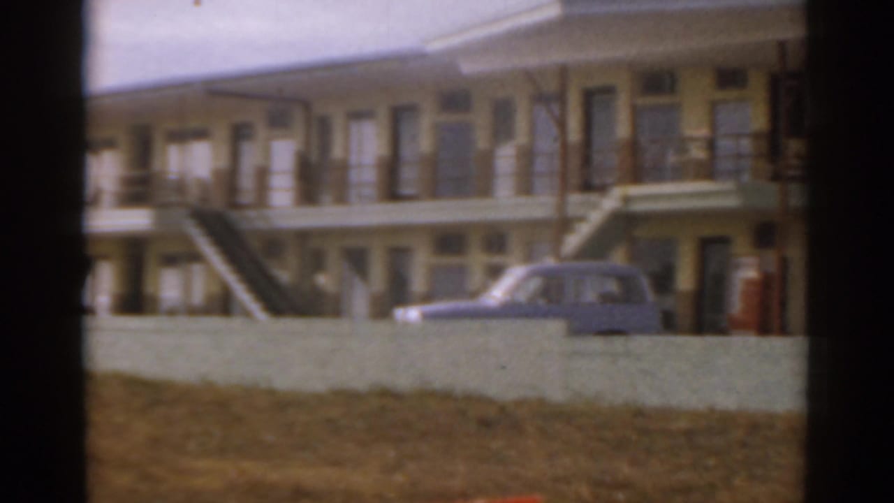 Yellow building with balconies and a blue car partly visible behind a low wall.