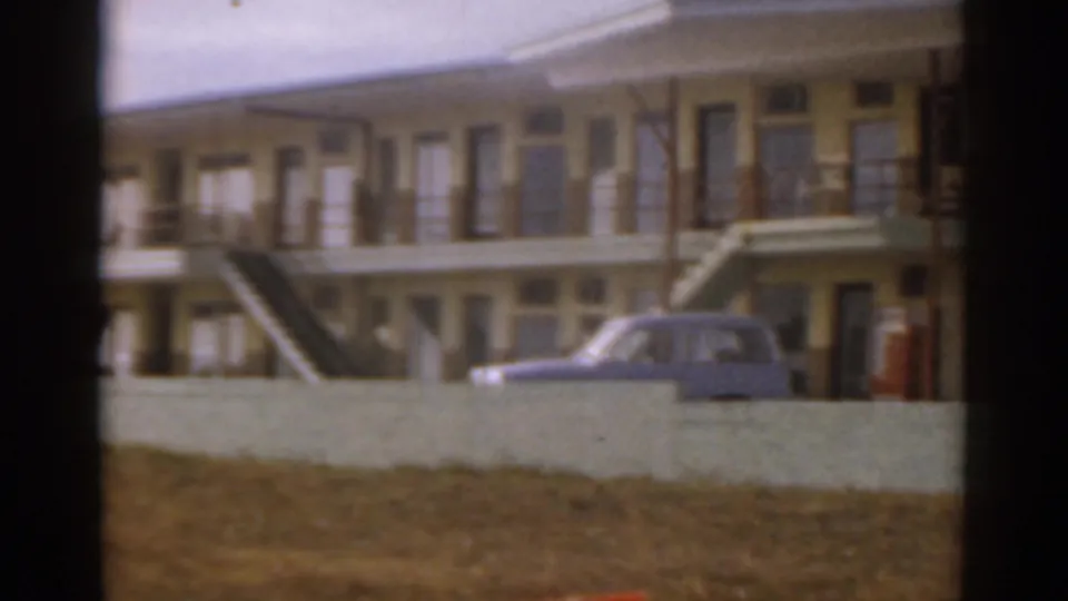 1963 Florida home-movie frame of a yellow beachfront lodging exterior, balcony walkway, exterior stair, and blue car near the wall.