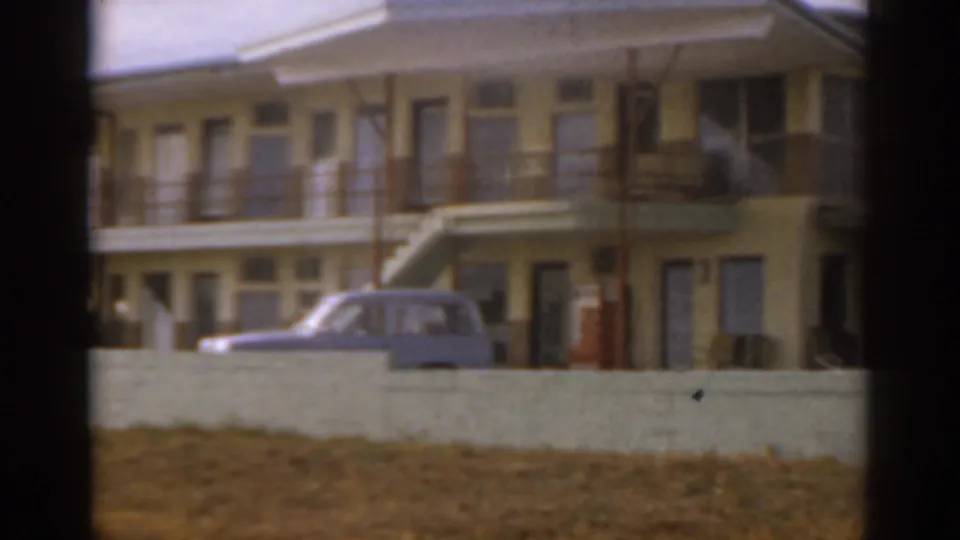1963 Florida 8mm frame showing a closer roadside view of the yellow beachfront building, blue car, balconies, and stairs.