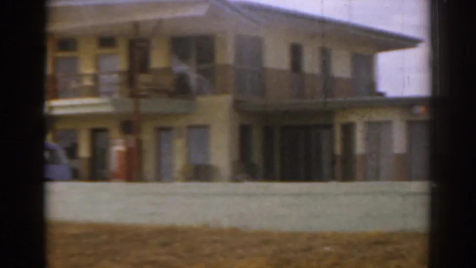 1963 Florida home-movie frame of the yellow building facade, low wall, exterior walkways, and partial blue car at frame edge.