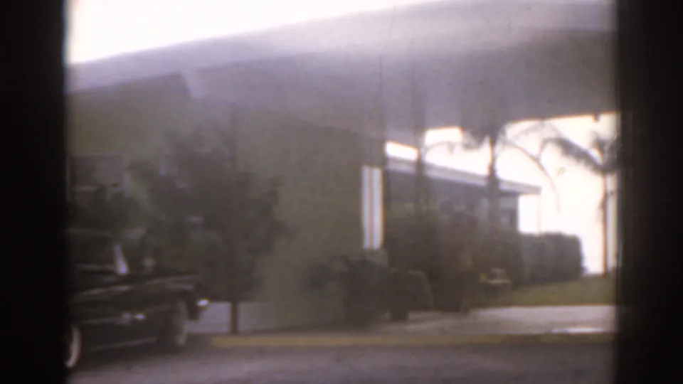 1963 Florida 8mm frame showing a rainy street, parked car, palms, shrubs, and a bright roadside building.
