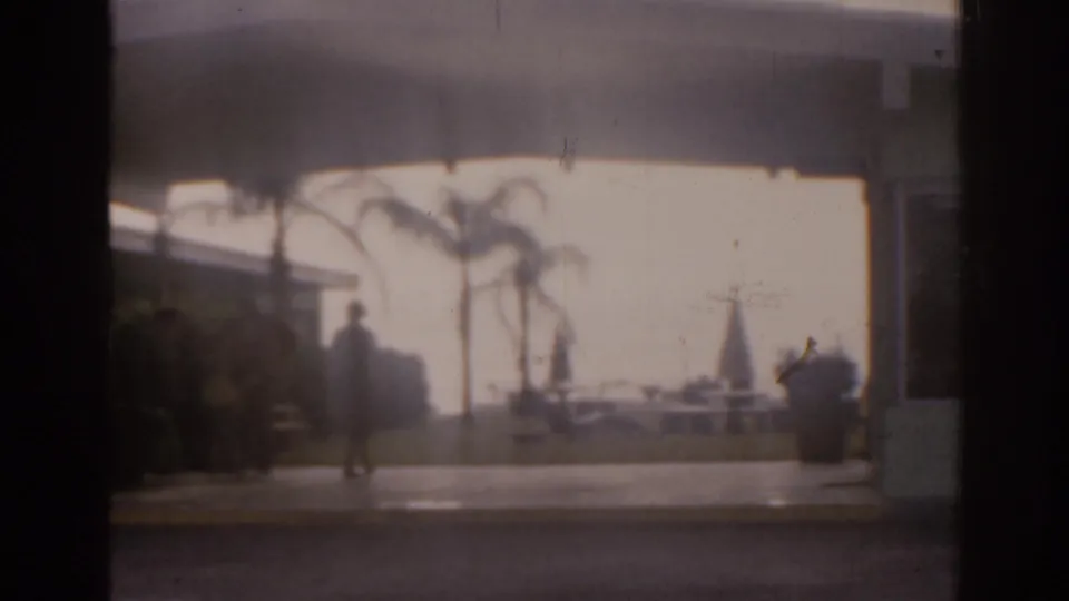 1963 Florida 8mm frame showing a soft, shaded view toward a low building entrance, palm trees, outdoor furniture, and a distant pedestrian.