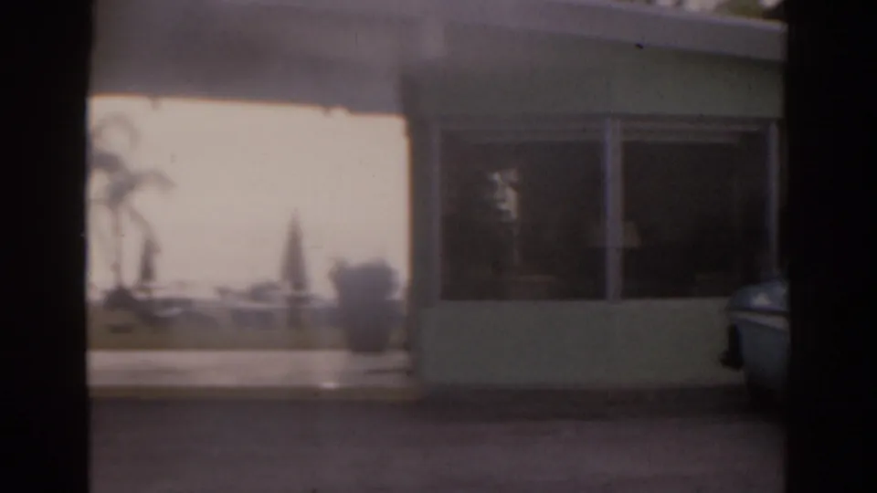 Soft 1963 Florida frame showing the side of a low building, a large dark window, palms, outdoor seating, and a partial parked car.