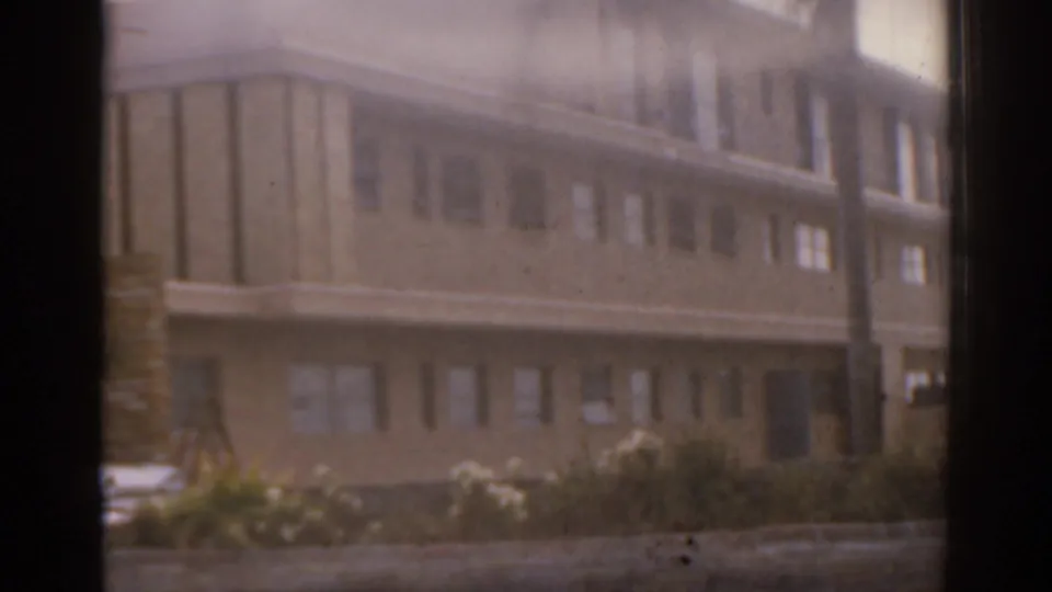 1963 Florida 8mm frame showing a brown multi-story building facade with repetitive windows, palms, shrubs, and partially visible parked cars.