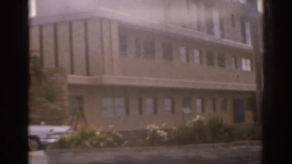 1963 Florida 8mm frame showing a multi-story building, vertical window panels, low shrubs, and cars in a parking lot.