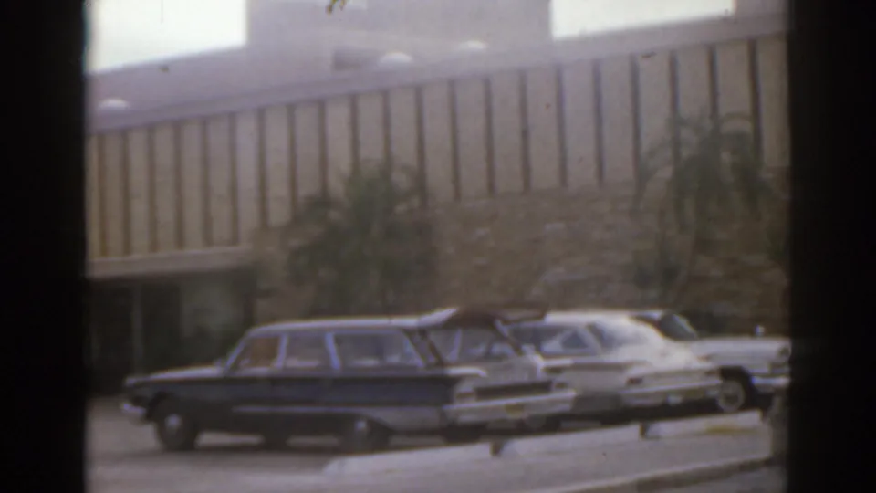 1963 Florida frame showing a dark station wagon, white sedan, palms, and a mid-century building exterior.