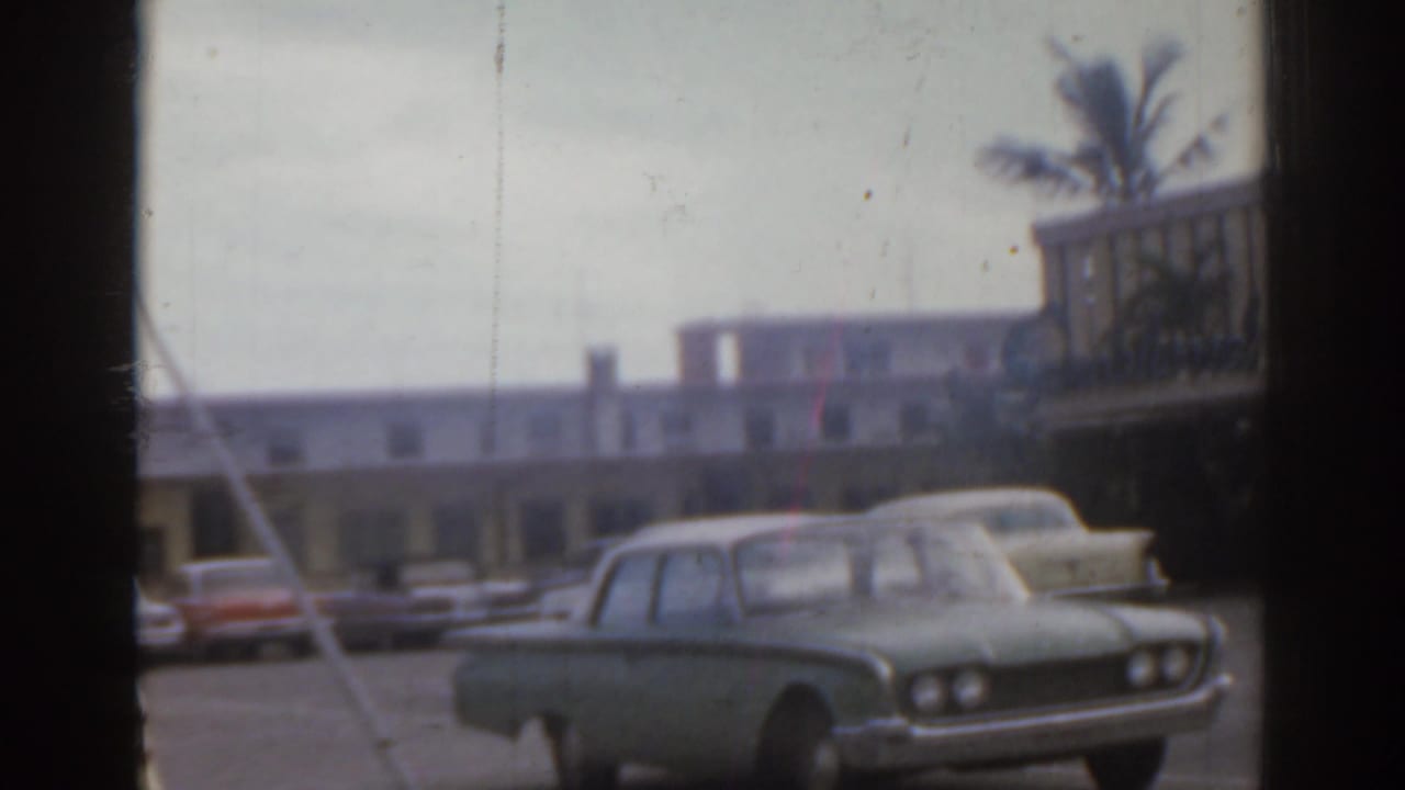 Parked 1960s cars outside a low roadside building, seen through dark vertical frame bars.