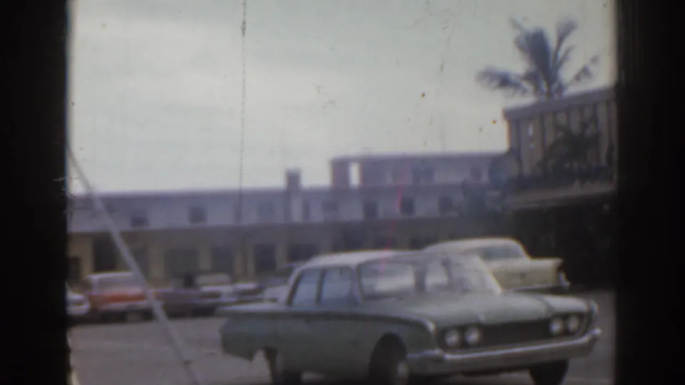 1963 Florida home-movie view of parked mid-century cars near a motel-like roadside building, partially blocked by window or vehicle framing.