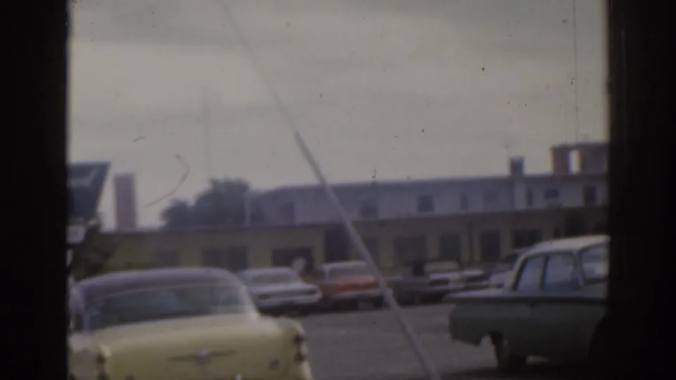 A yellow sedan and other 1960s cars appear in a Florida roadside parking area, seen through obstructing window or vehicle frame bars.