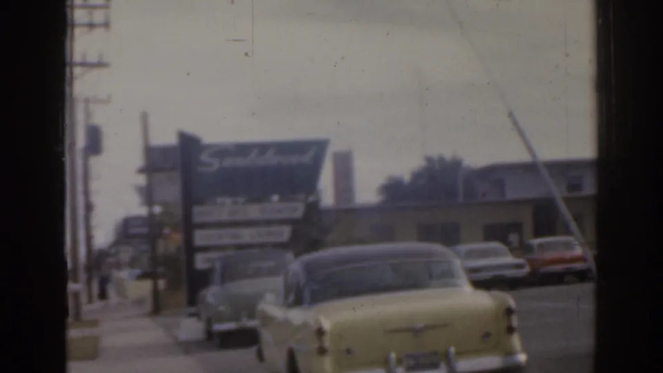 1963 Florida street view with a large motel-style sign, parked mid-century cars, utility poles, and sidewalk in soft 8mm focus.