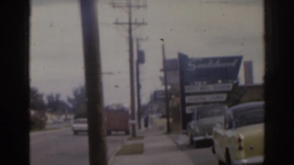 A Florida roadside sign and parked 1960s cars line a commercial street, with utility poles and soft home-movie film texture visible.