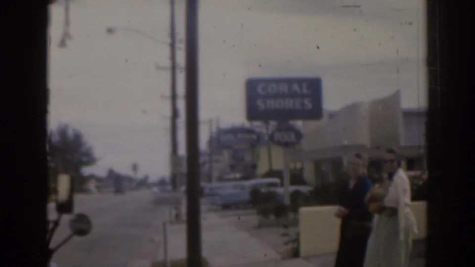 1963 Florida home-movie frame showing two women by a roadside Coral Shores sign with cars and motel buildings behind them.