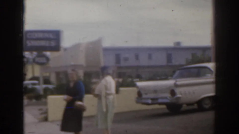 1963 Florida frame showing two women near a motel frontage as a white tailfin-era station wagon passes in the street.