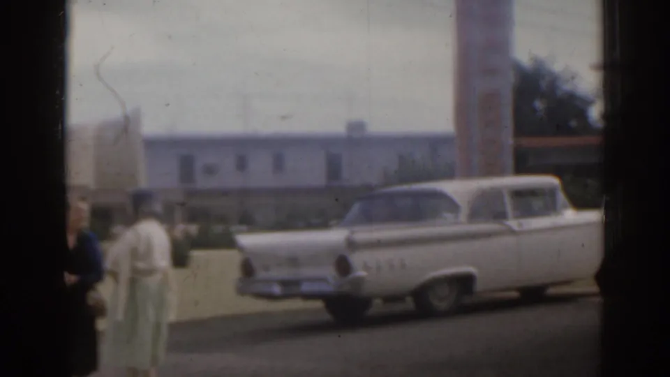 1963 Florida home-movie frame with a white station wagon in front of motel buildings and two women partly visible by a wall.
