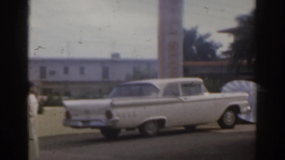 1963 Florida home-movie frame showing a white American car parked near motel buildings, partly obscured by dark window bars.