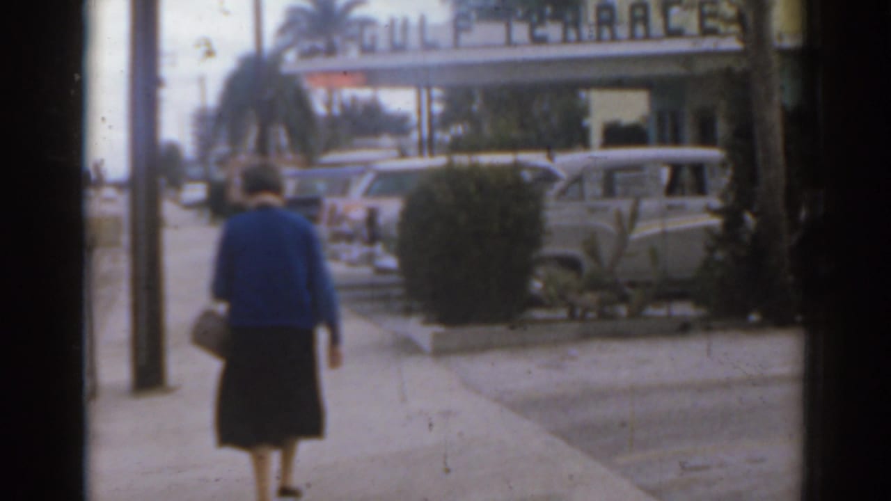Woman in a blue sweater walks away on a sidewalk beside parked cars.