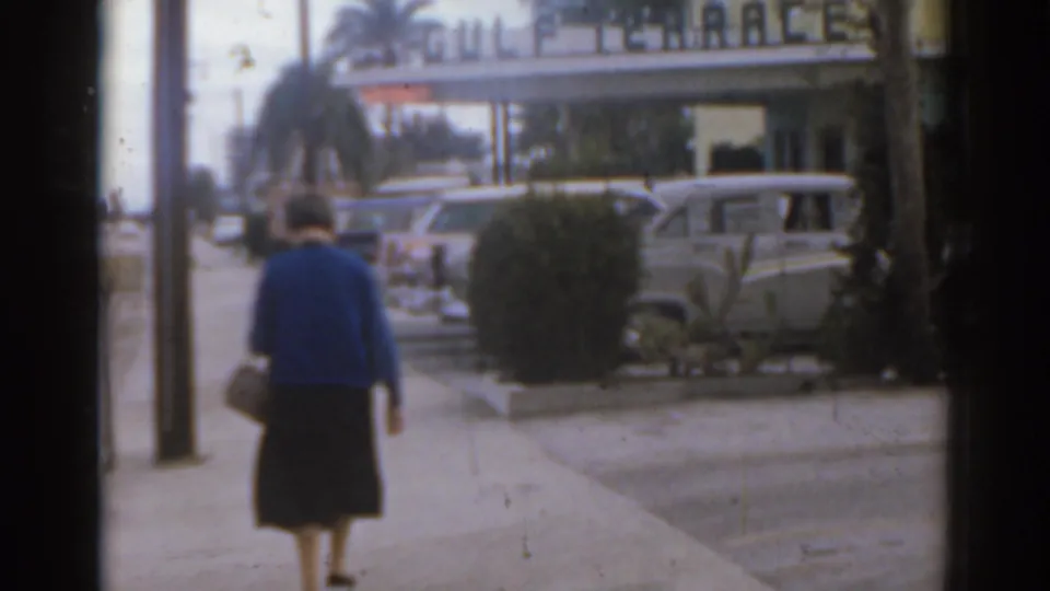 1963 Florida home-movie frame showing a woman walking away on a sidewalk near parked cars and a Gulf Terrace sign.