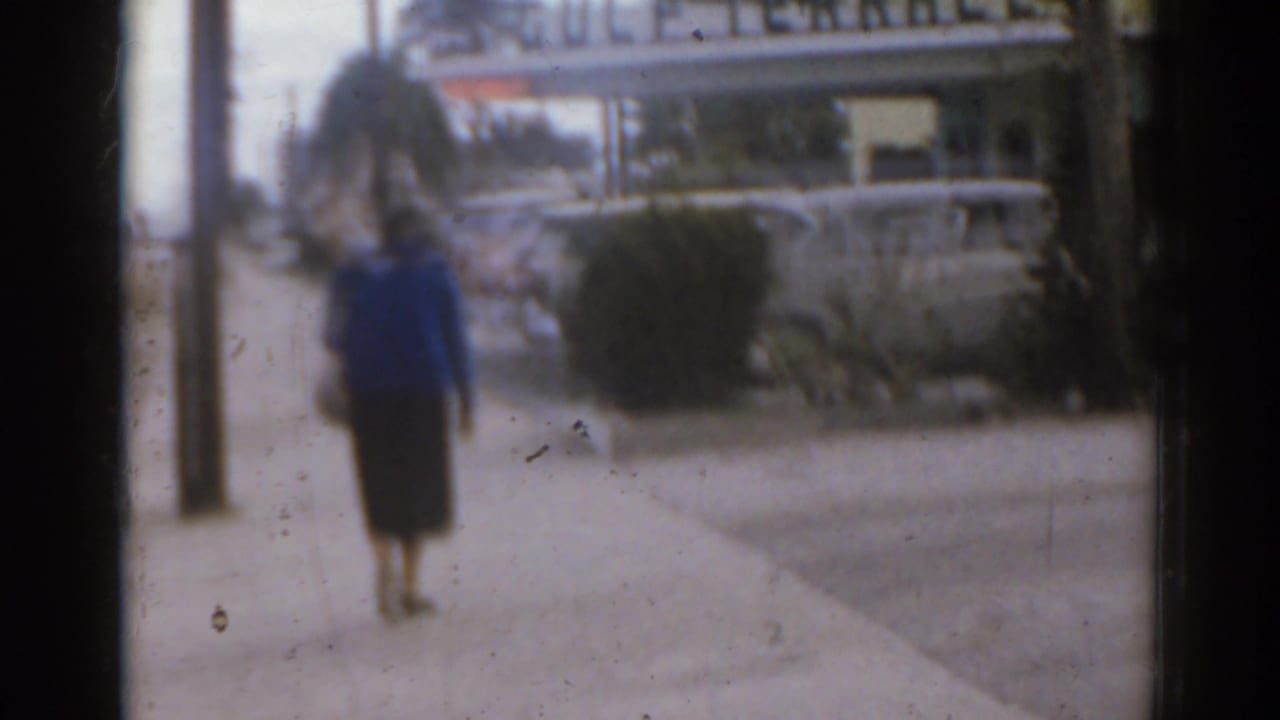 Woman stands or walks on a sidewalk with parked cars and a sign behind her.