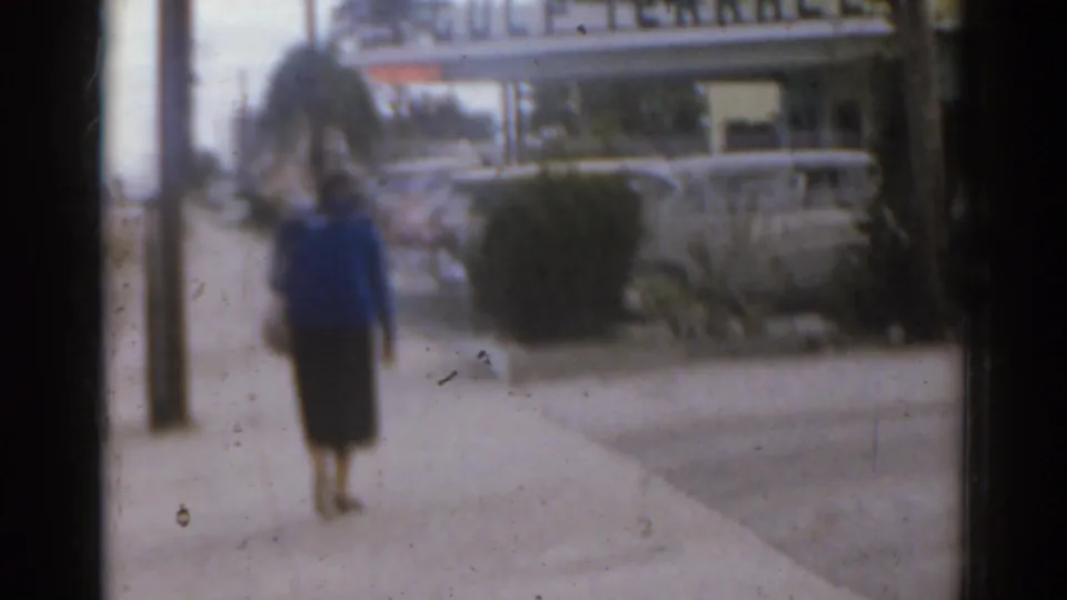 Soft 1963 Florida frame of a woman on a sidewalk, with palm trees, parked cars, and Gulf Terrace signage in the background.