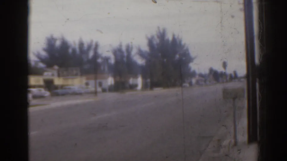 1963 Florida street view showing a roadway, trees, a yellow building, and a decorative gate in soft 8mm focus.