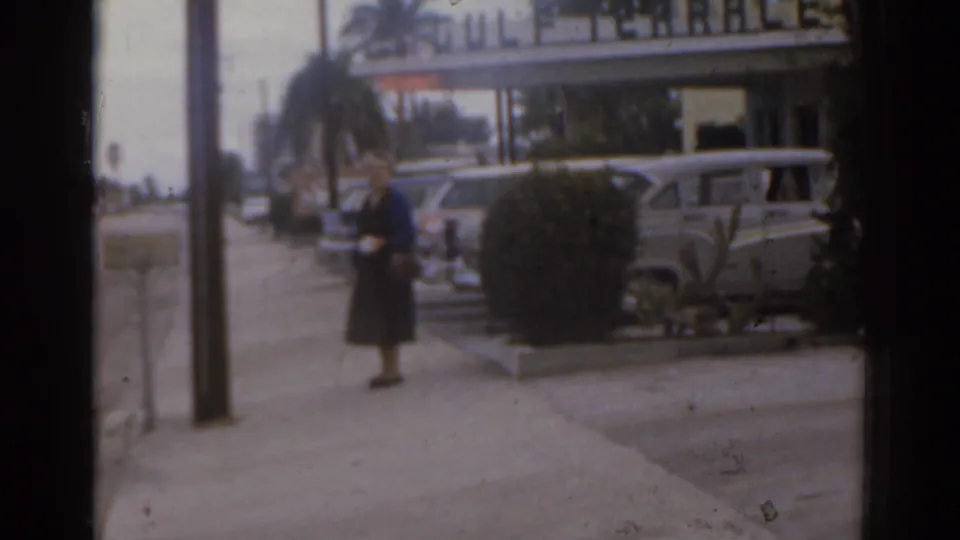 1963 Florida home-movie frame of a woman on the sidewalk near a utility pole, parked cars, palm trees, and Gulf Terrace signage.