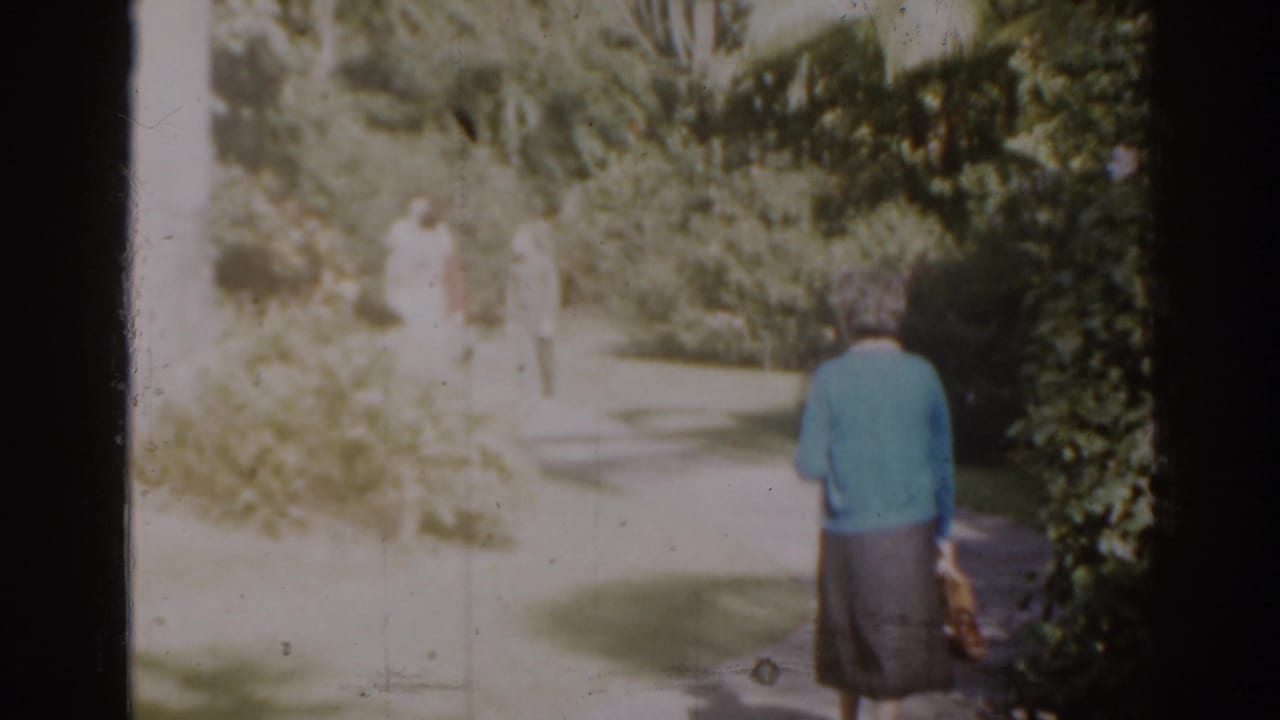 Older woman in a blue sweater walking along a garden path with distant figures ahead.