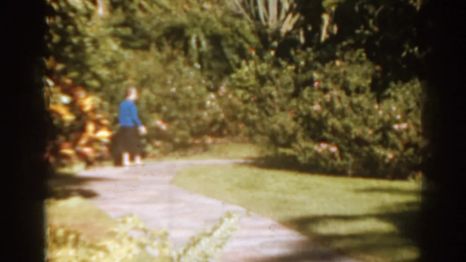 In 1963 Florida footage, a woman in a blue sweater walks away along a sunlit garden path bordered by shrubs.