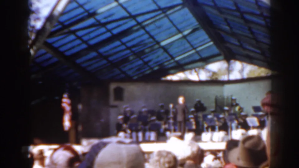 The band continues under the glass roof, seen from behind spectators, with a partial face visible at the right edge of the frame.