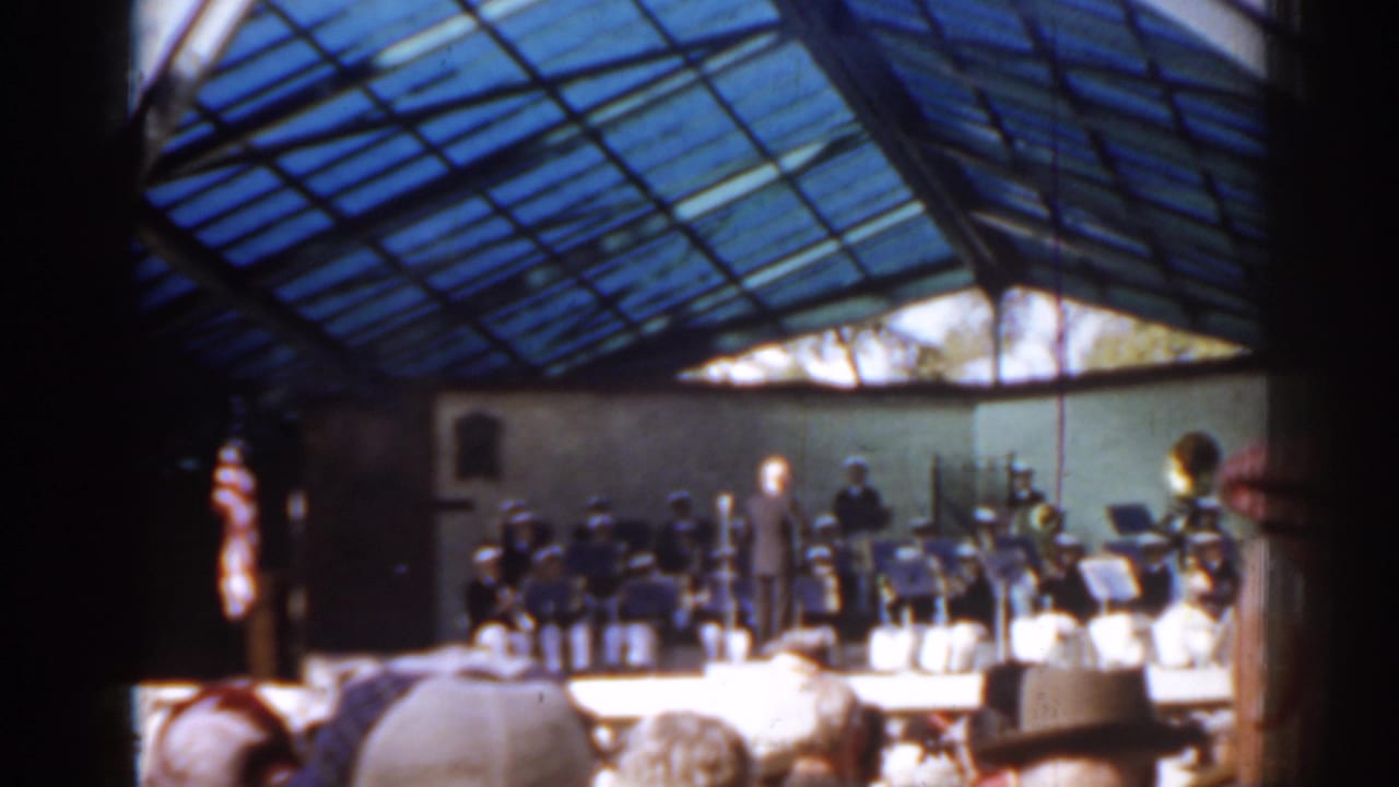 Band performing under a blue glass roof while an audience watches from the foreground.