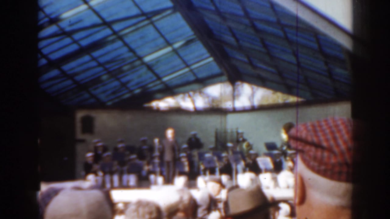 1963 Florida home-movie view of a public concert, with a conductor centered on stage and audience hats framing the foreground.