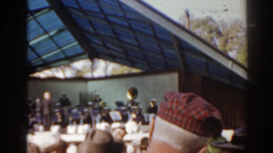 Late-frame 1963 Florida concert view with band, conductor, audience hats, and the venue's blue-tinted canopy visible.