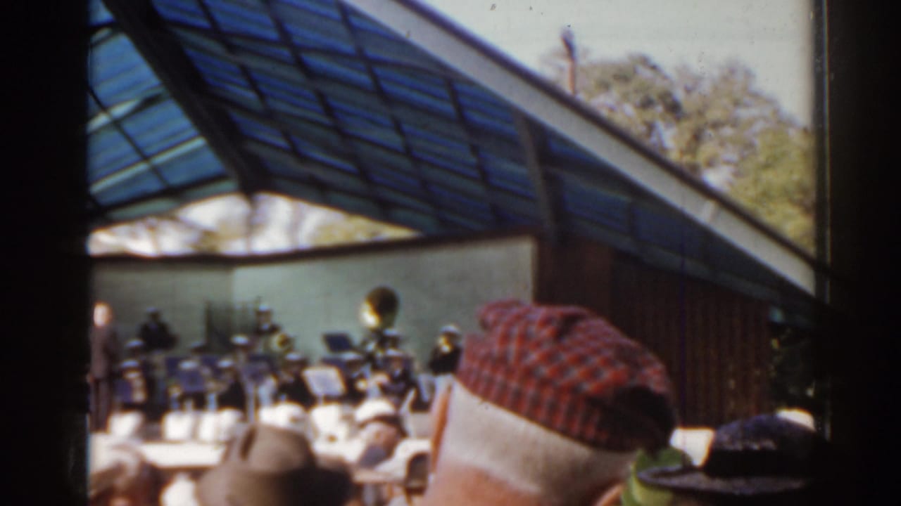 Spectators sit and stand near a band under a glass-roofed pavilion.