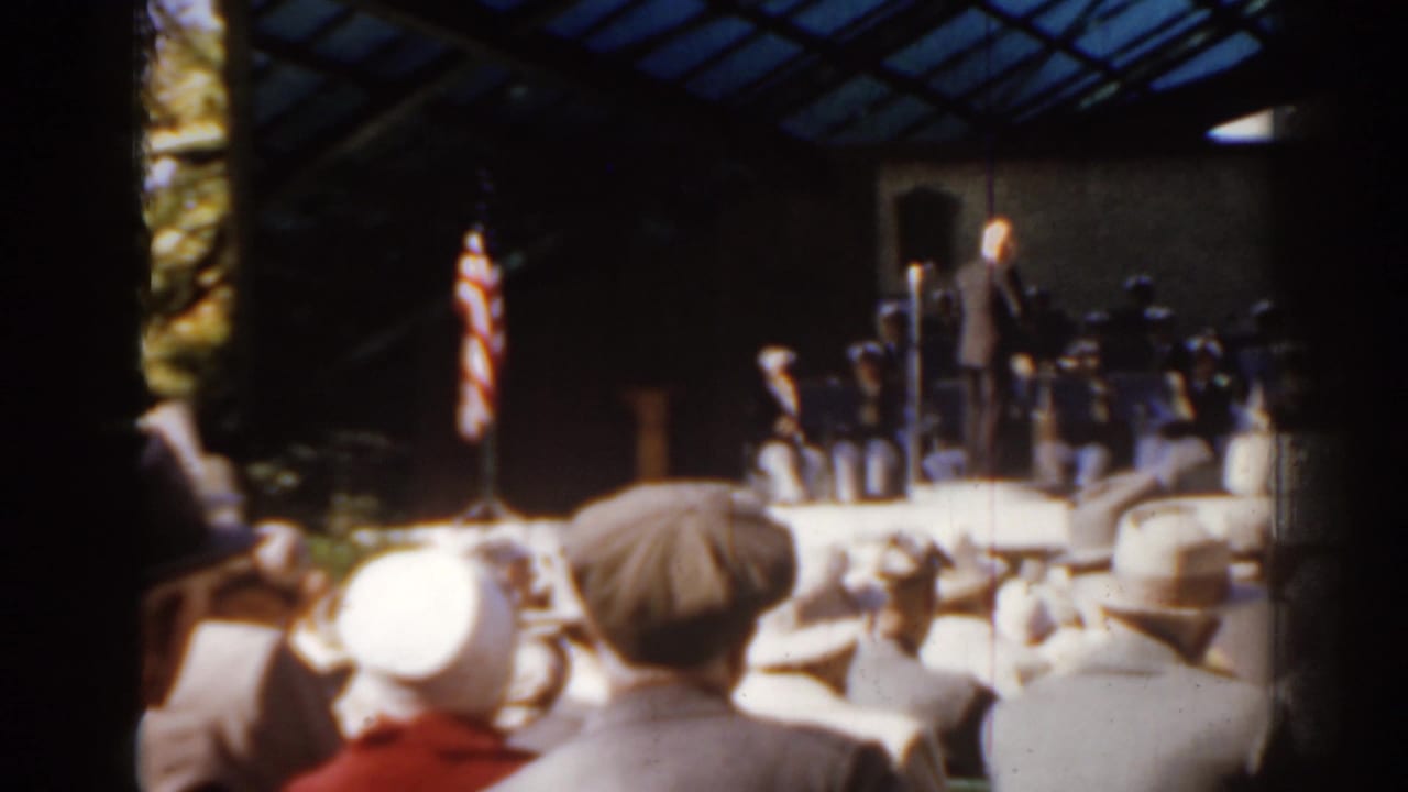 Conductor stands before a band as spectators watch from the grass.