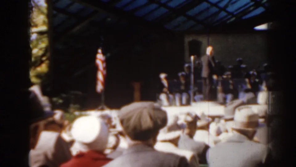 1963 Florida frame with a conductor leading musicians on a raised outdoor band area while spectators sit in the foreground.