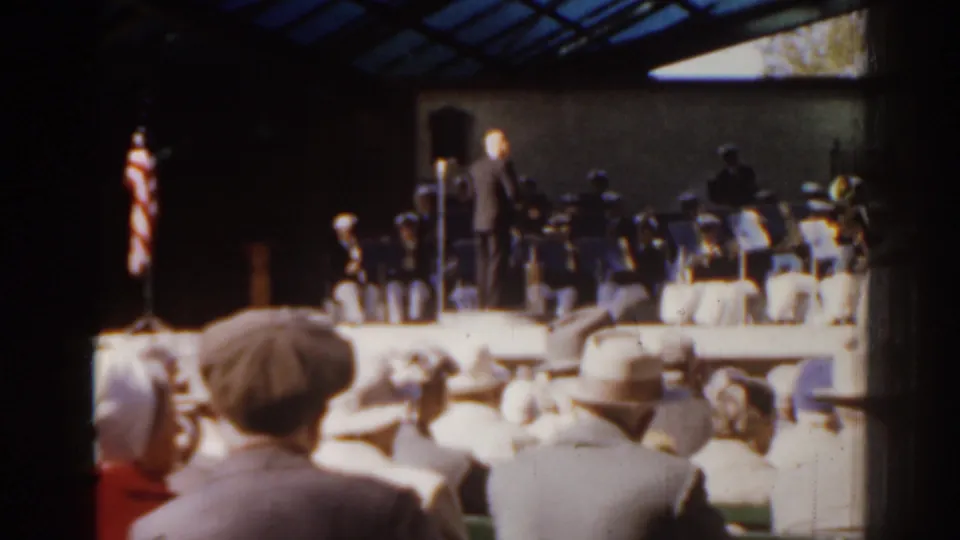 1963 Florida frame showing spectators in period clothing watching a conductor lead musicians beneath a glass-roofed pavilion.
