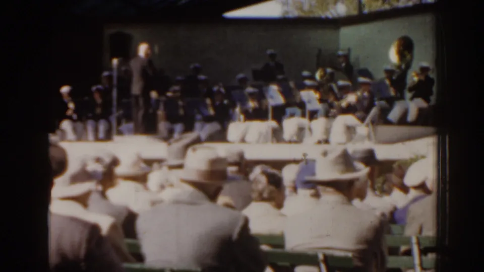 A soft 8mm frame shows a conductor leading musicians as a public audience watches from benches in Florida.
