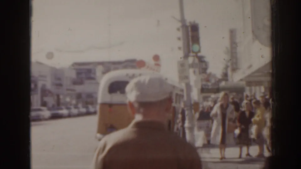 1963 Florida 8mm frame showing a yellow-and-white bus, pedestrians, traffic lights, and a sidewalk viewed from behind a passerby.
