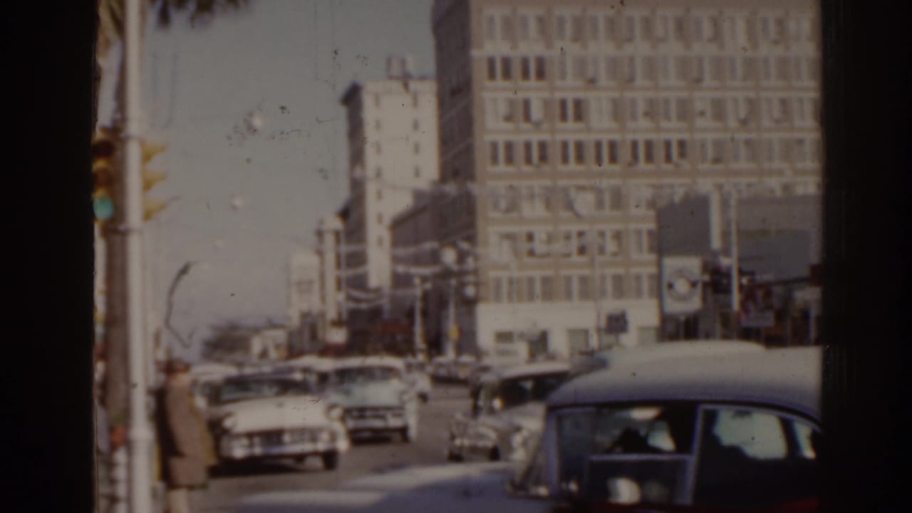 Classic cars pass through an intersection beside tall city buildings.
