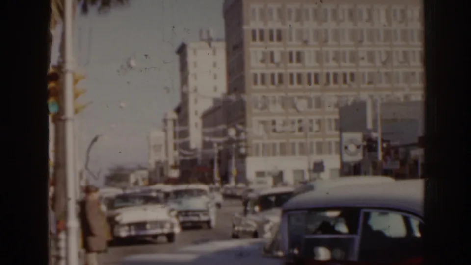 1963 Florida street view with classic cars, traffic signals, pedestrians, and a tall gridded building in the background.