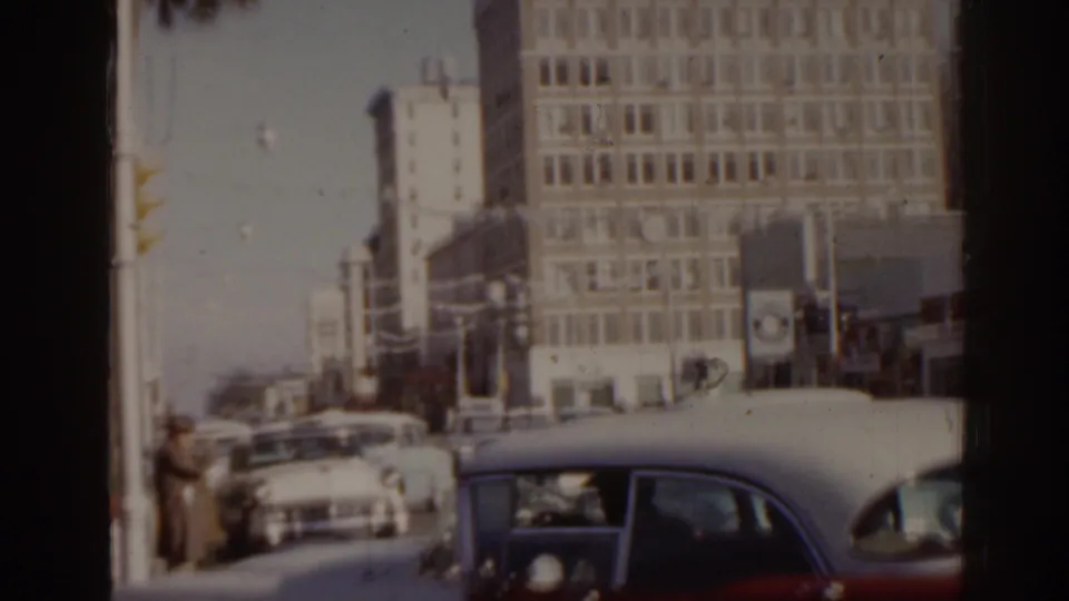 1963 Florida 8mm frame with classic cars, a dark foreground car, pedestrians, traffic lights, and multi-story commercial buildings.