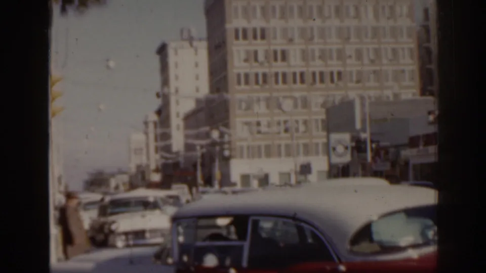 1963 Florida city street frame showing moving classic cars, pedestrians near the curb, traffic lights, and a large gridded building.