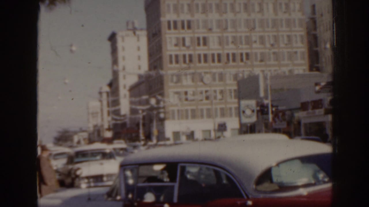 Downtown street with classic cars, pedestrians, traffic lights, and mid-century buildings.