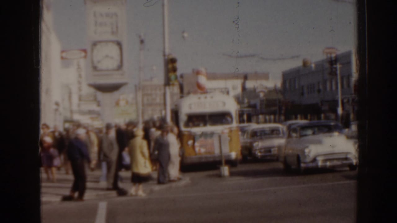 1963 Florida intersection with pedestrians crossing, classic cars, a white-and-yellow bus, traffic lights, and a large street clock.