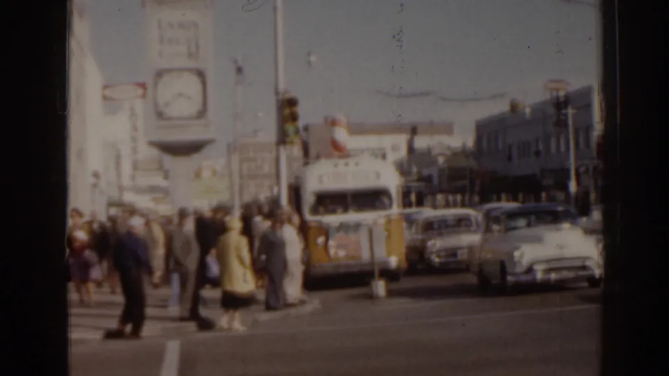 1963 Florida intersection with pedestrians crossing, classic cars, a white-and-yellow bus, traffic lights, and a large street clock.