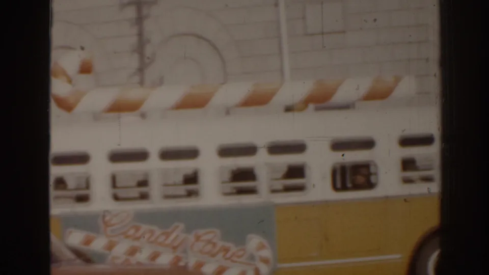 A close side view of the candy-cane-decorated bus in 1963 Florida, with windows, yellow panels, and a pale building behind it.