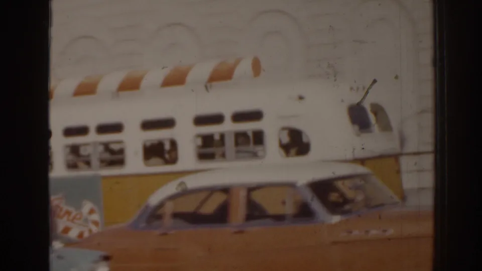 1963 Florida street view with the decorated bus partly behind a tan classic car, showing traffic and storefront-scale architecture.