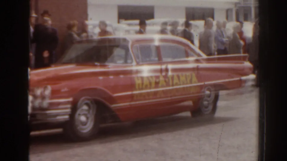 1963 Florida 8mm frame showing the side of a red Hav-A-Tampa car with a sidewalk crowd in the background.
