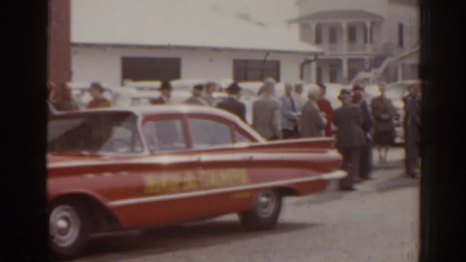1963 Florida frame showing the rear side of a Hav-A-Tampa car and adults gathered near brick and white buildings.