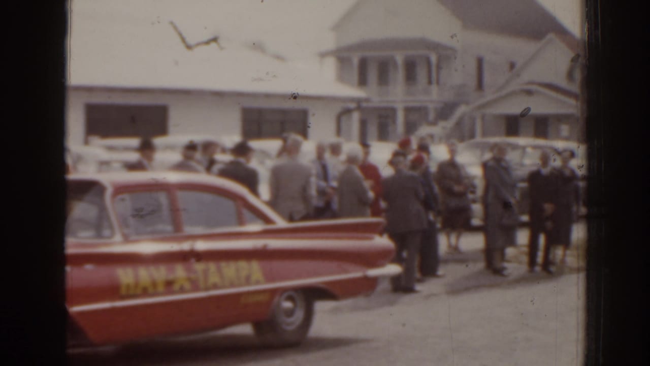 Uniformed adults stand on a street beside a red HAV-A-TAMPA car with houses behind them.