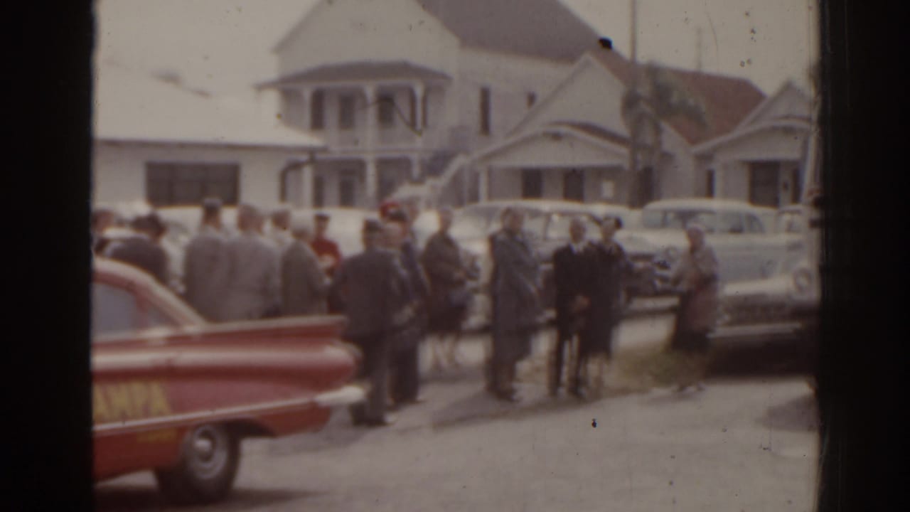 Grey-uniformed adults stand in a Florida street scene with a red lettered car and mid-century parked sedans.